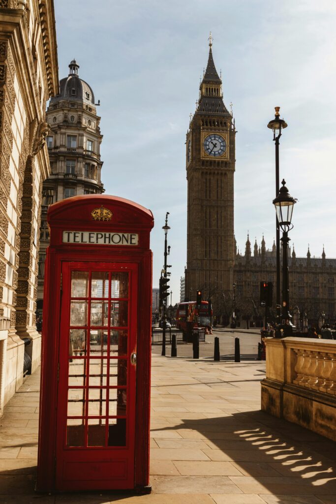 Red telephone booth with Big Ben and historic London architecture in daylight.